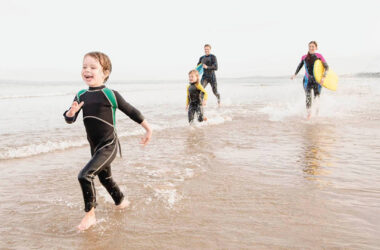 Family enjoying beach with wetsuits, splashing in sea near hotel.