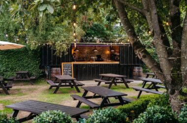 Charming garden bar with picnic tables, lush greenery, and string lights creating a cosy, inviting atmosphere for relaxation.