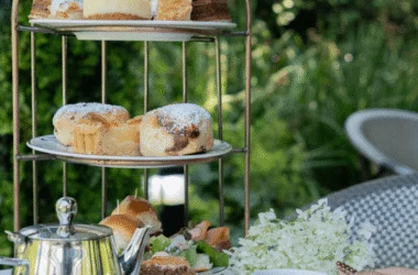 Afternoon tea in serene garden with tiered cake stand, scones, pastries, and silver teapot, offering a relaxing experience.