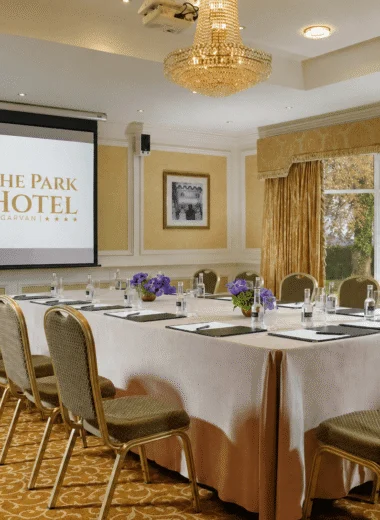 Conferencing set up with a screne for a presenation with the park hotel logo in it with a big table in front with a number of seats and water and note paper on the table