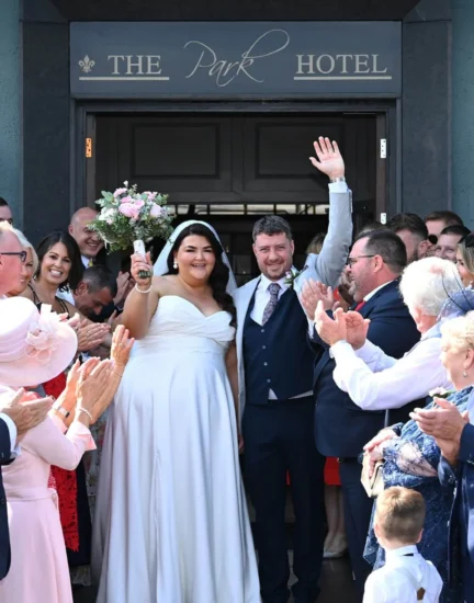 Smiling wedding couple exit Park Hotel surrounded by cheering guests and flowers, showcasing joyful celebration venue.